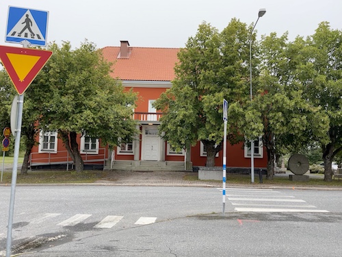 Tien toisella puolella punainen talo ja edessä vihreitä puita.